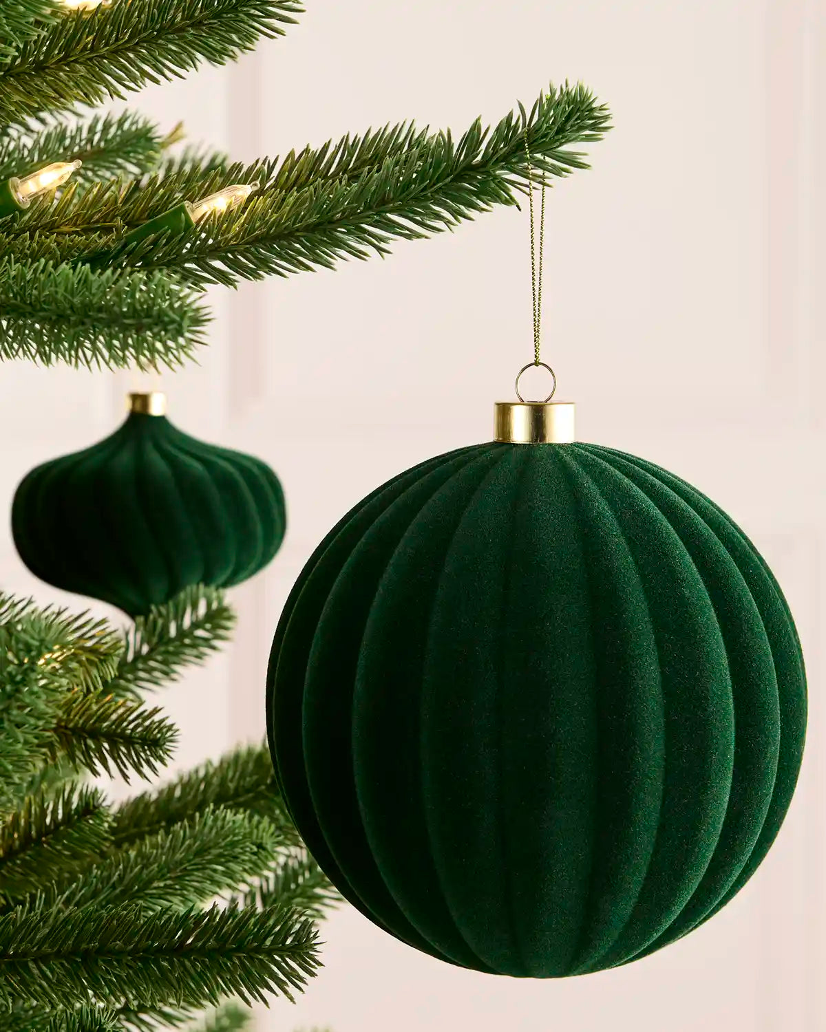 A sage green ribbed velvet Christmas ornament hanging from a green Christmas tree branch, with a second matching ornament blurred in the background.