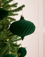 A sage green ribbed velvet Christmas ornament hanging from a green Christmas tree with warm white string lights, against a soft off-white paneled wall background.