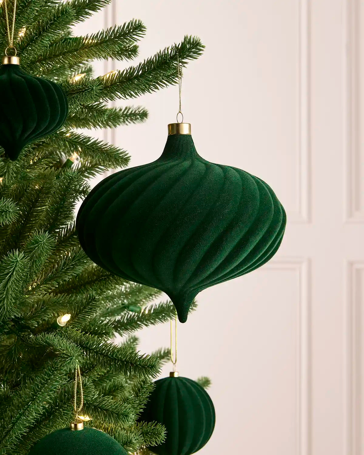 A sage green ribbed velvet Christmas ornament hanging from a green Christmas tree with warm white string lights, against a soft off-white paneled wall background.