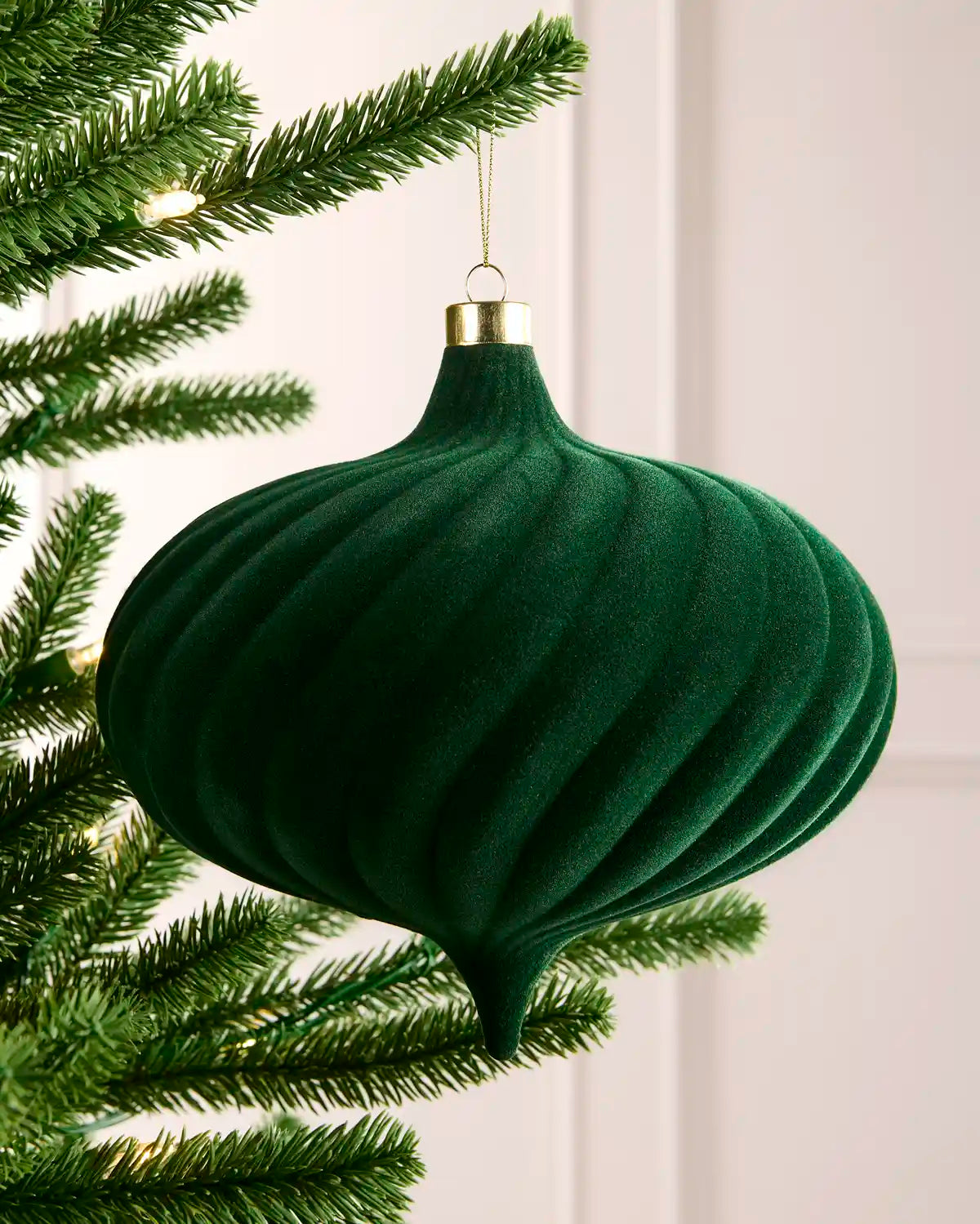 A sage green ribbed velvet Christmas ornament hanging from a green artificial tree branch against a soft off-white paneled background.