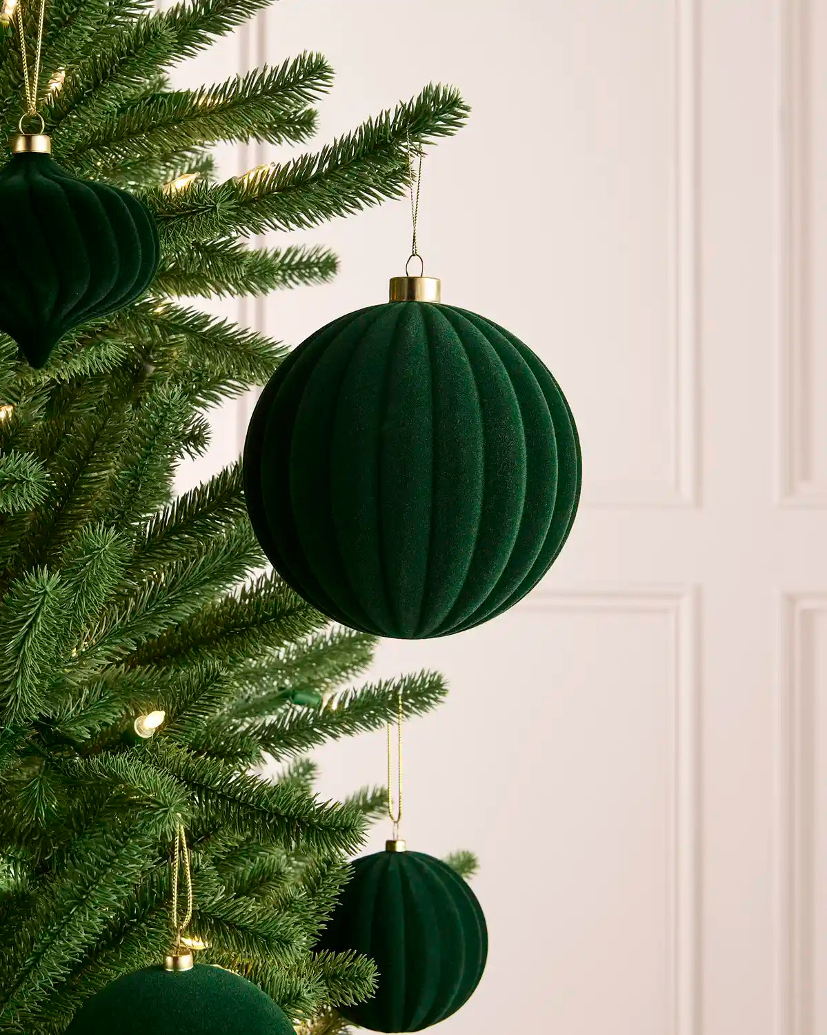 A sage green ribbed velvet Christmas tree ornament hanging from a green artificial Christmas tree, with a soft cream paneled wall in the background.
