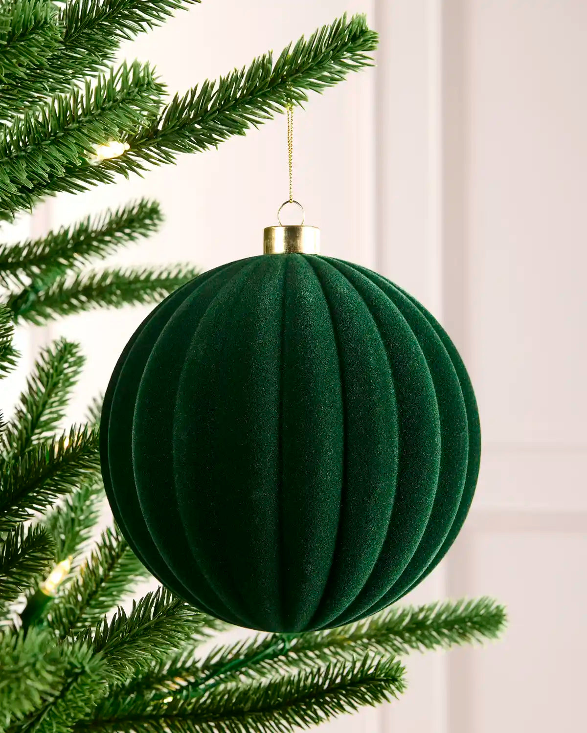 A sage green ribbed velvet Christmas tree ornament hanging from a green artificial tree branch with warm string lights in the background.