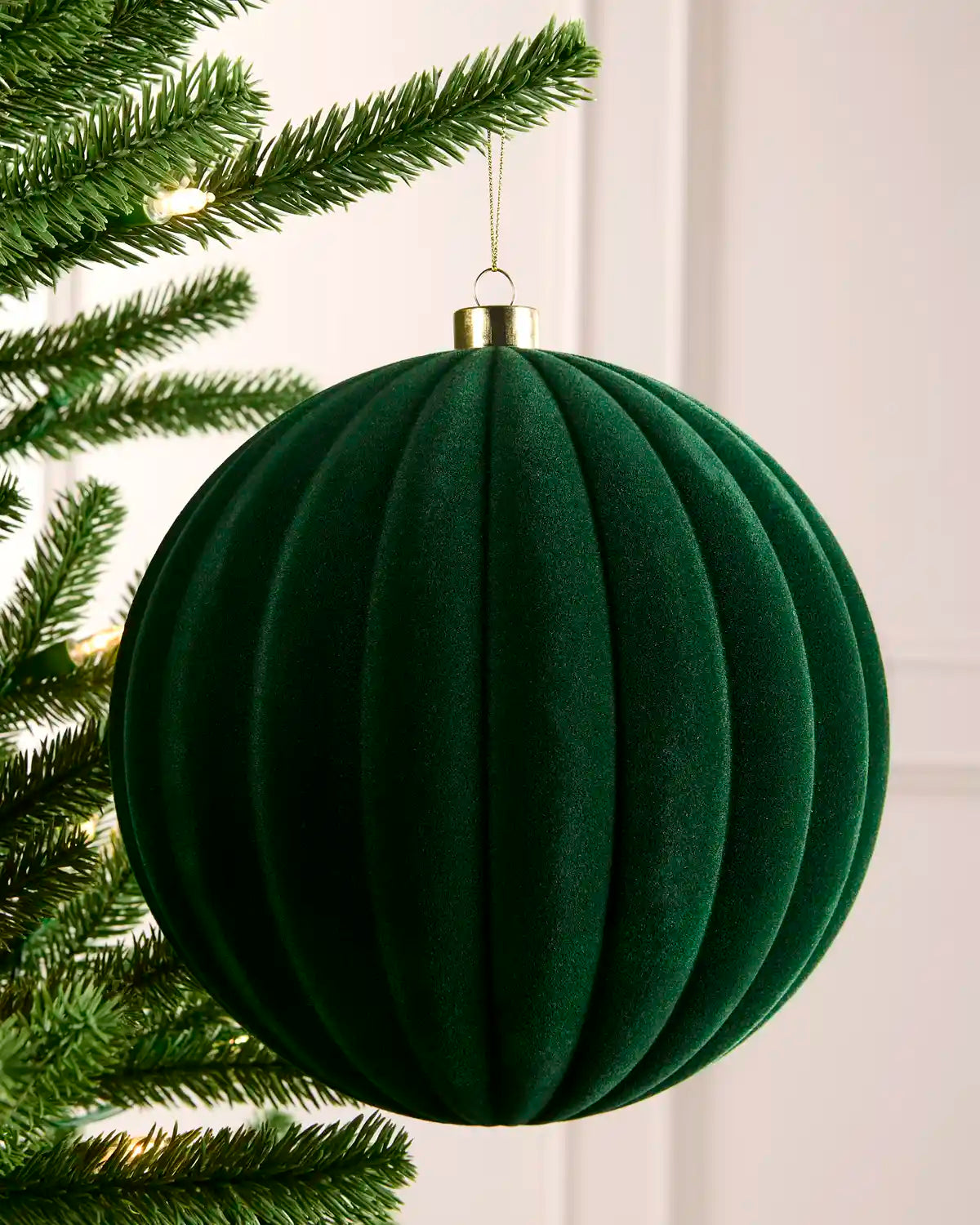 A sage green ribbed velvet Christmas tree ornament hanging from a green pine tree branch with warm string lights in the background.
