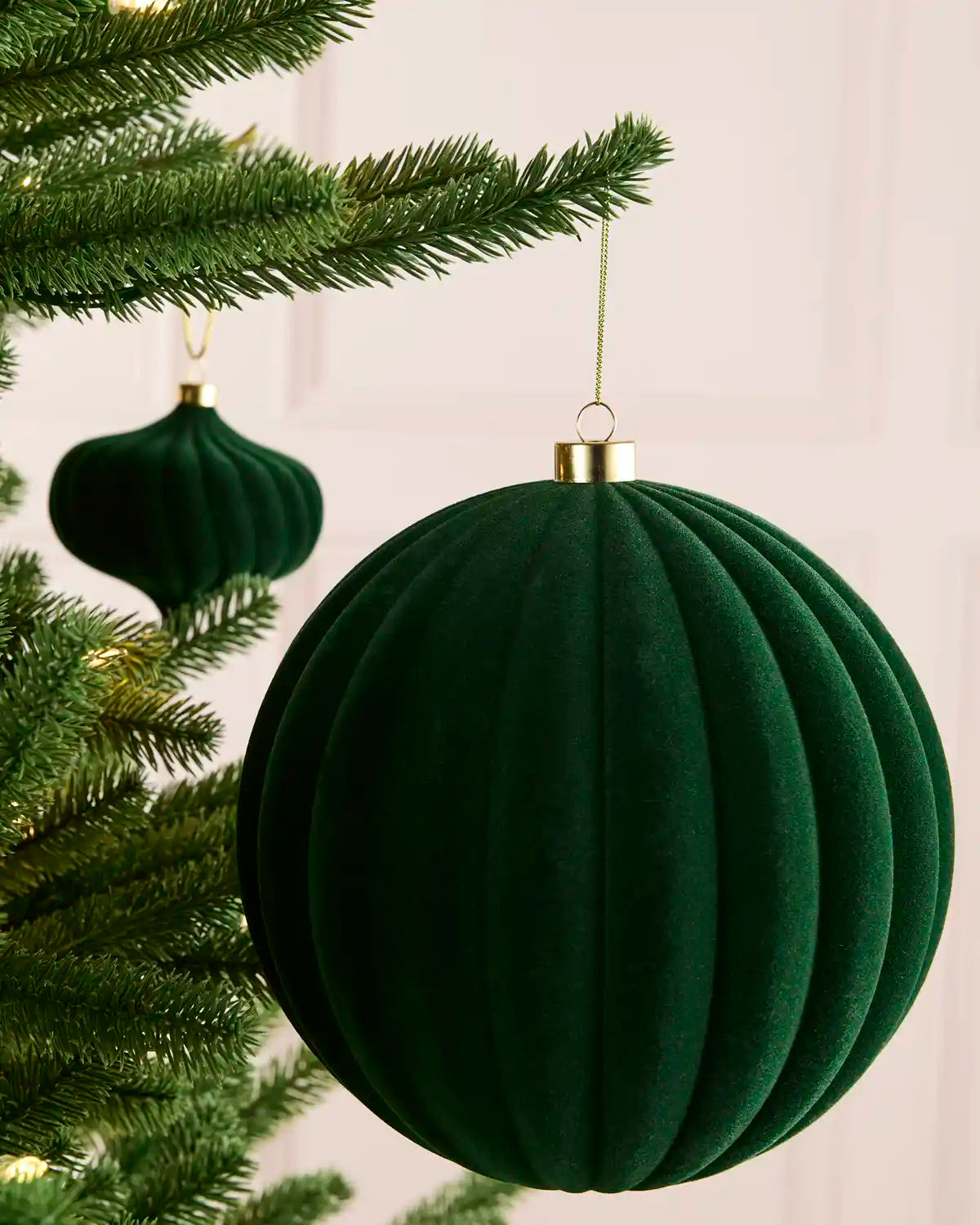 A sage green ribbed velvet Christmas tree ornament hanging from a pine tree branch, with a gold cap and a gold hanging string.