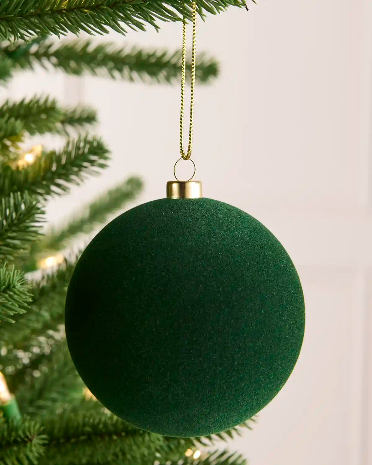 A sage green textured spherical Christmas ornament hanging from a gold wire hanger on a pine tree branch, with warm string lights softly blurred in the background.