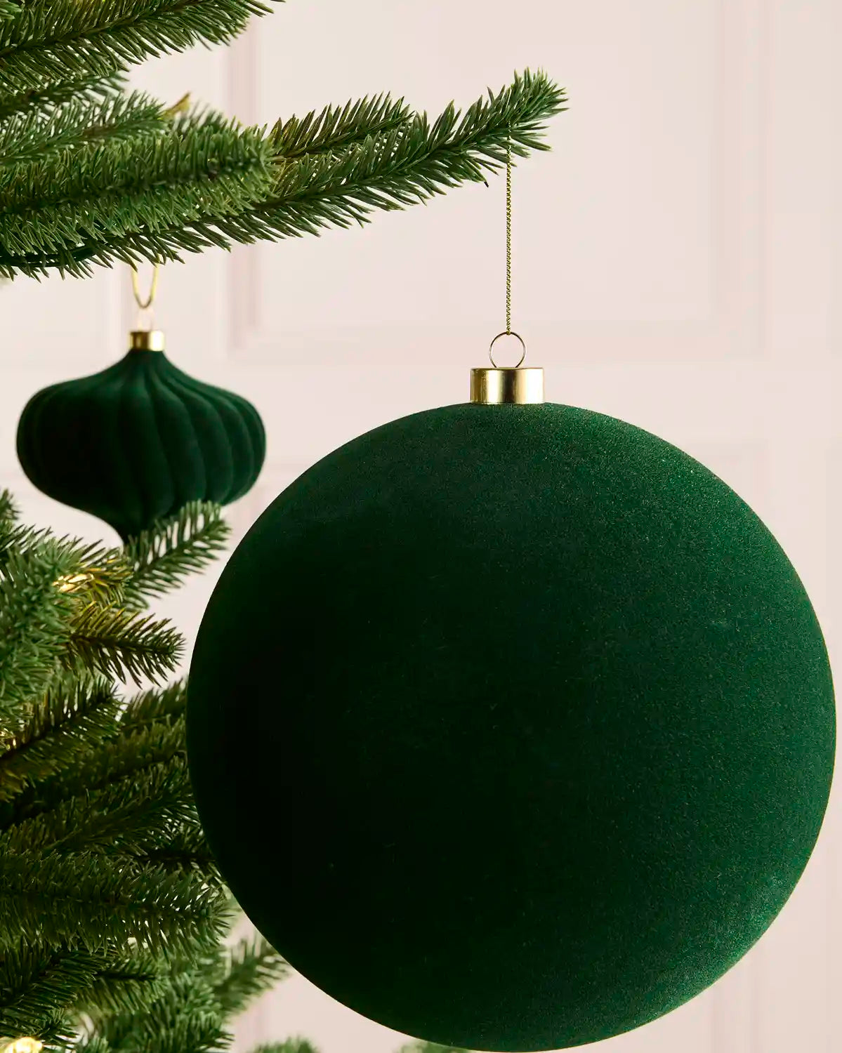 A sage green velvet Christmas bauble ornament hanging from a gold cap on a pine tree branch, with a second matching ornament blurred in the background.