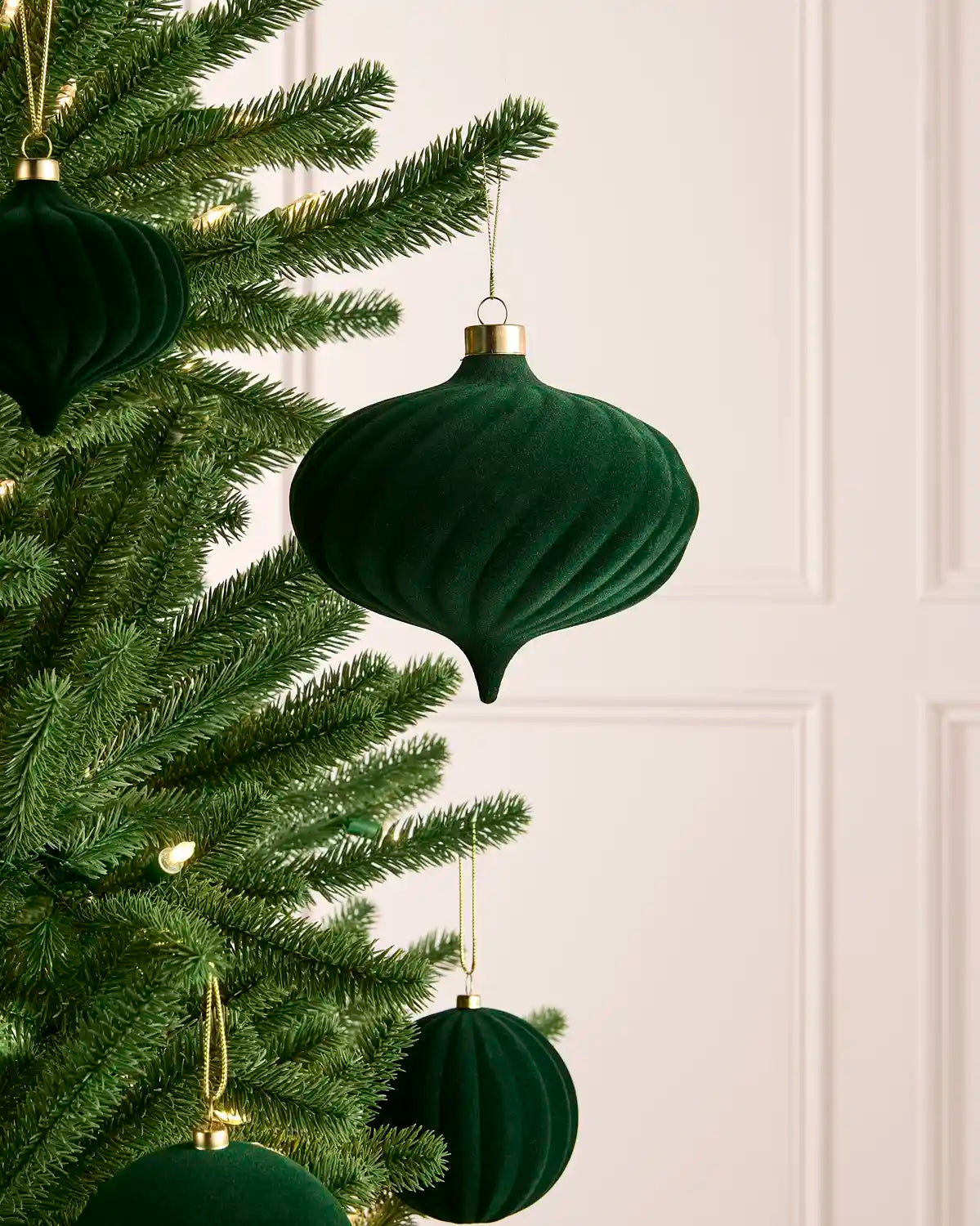 A sage green velvet Christmas ornament with a ribbed texture and gold cap, hanging from a green Christmas tree branch against a light cream paneled wall.