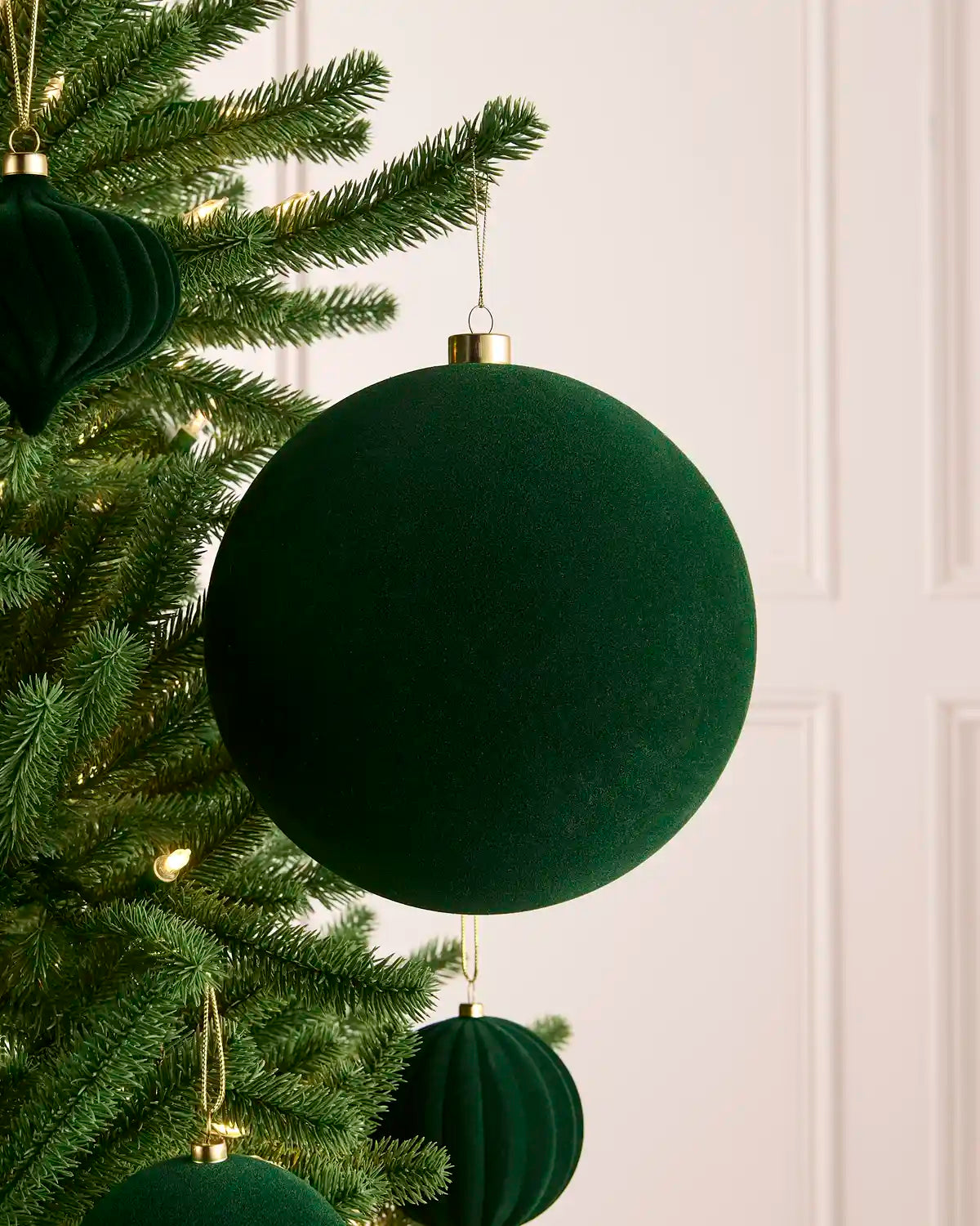 A sage green velvet Christmas tree ornament hanging from a green pine tree branch, with a soft-focus cream paneled wall in the background.