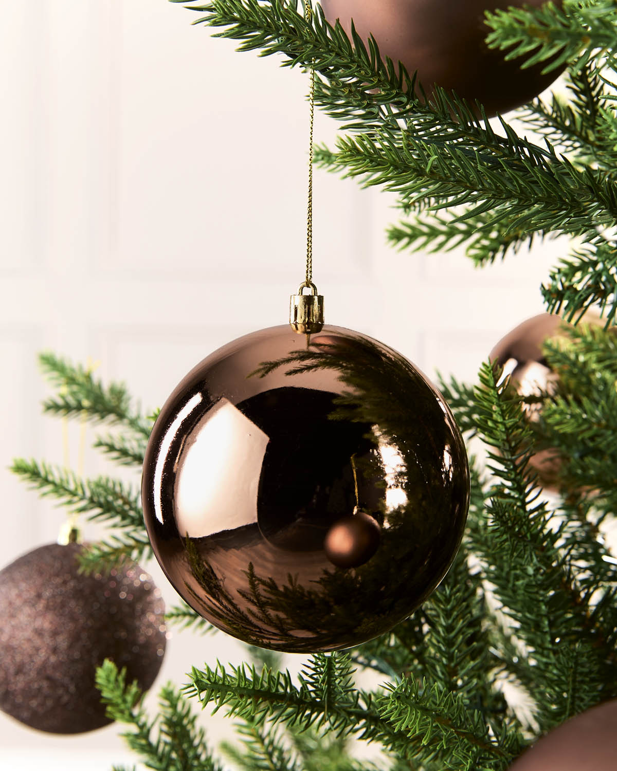 A shiny metallic espresso Christmas ornament hanging from a green pine tree branch, with other ornaments and the tree branches softly blurred in the background.