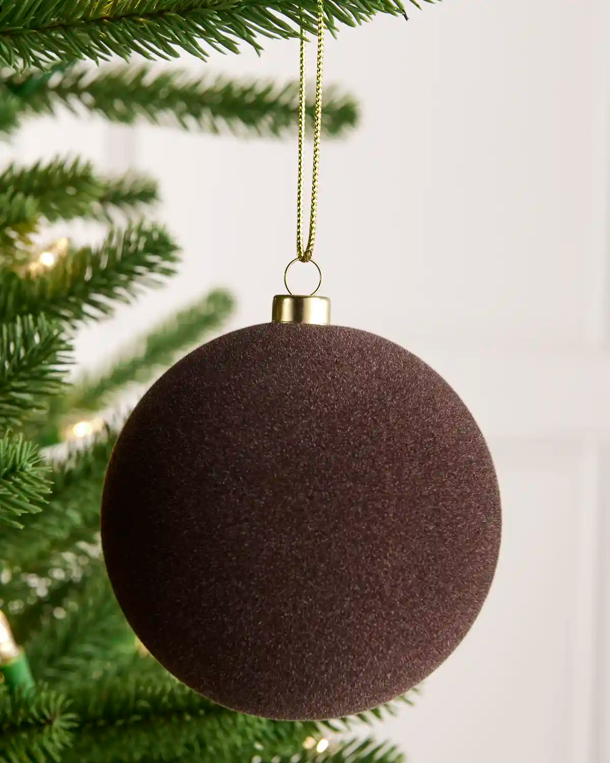 A single espresso coloured textured spherical Christmas ornament hanging from a gold string on a green Christmas tree branch against a white panelled background.