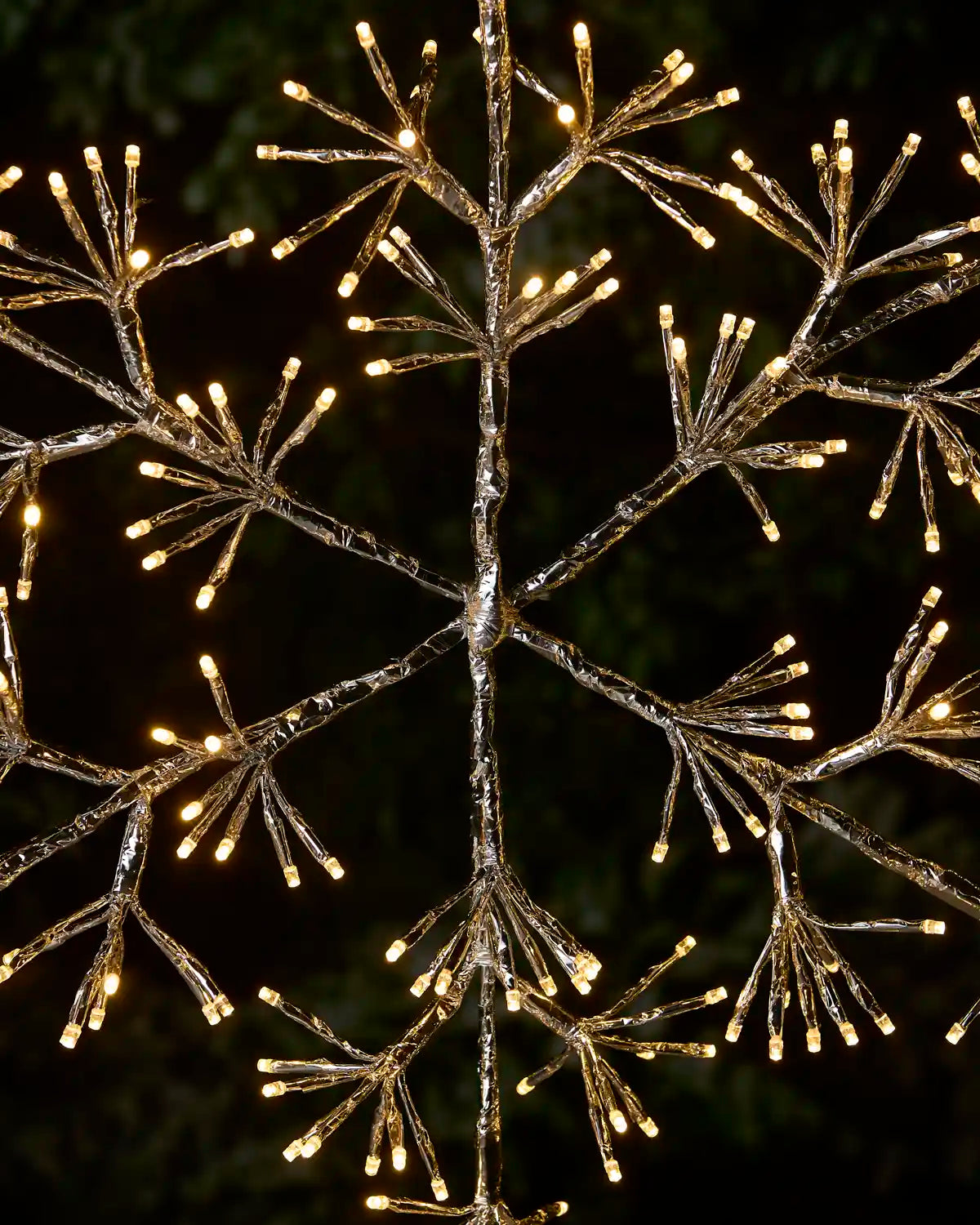 A snowflake-shaped decorative light fixture with warm white LED bulbs embedded in clear, branch-like plastic, shown against a dark outdoor background.