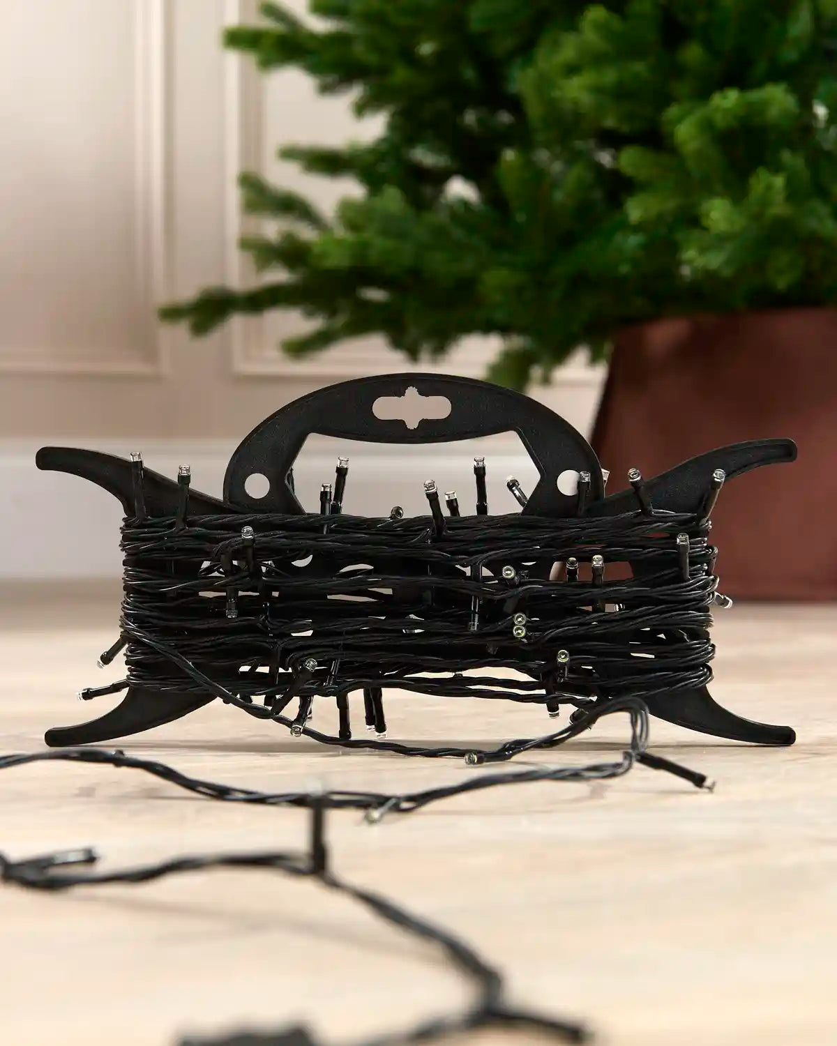 A spool of black Christmas string lights with small silver bulbs, displayed on a light wood floor with a blurred green Christmas tree in the background.