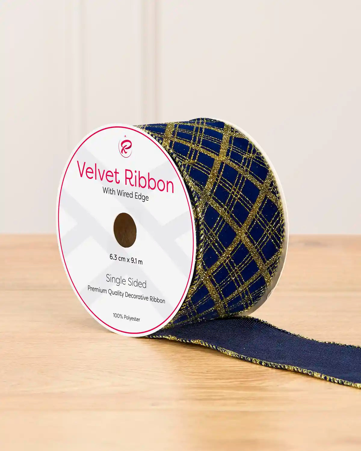A spool of velvet ribbon with a navy blue and gold plaid pattern, resting on a light wooden surface against a neutral beige background.