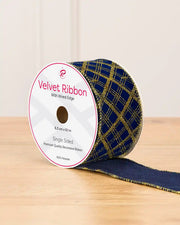 A spool of velvet ribbon with a navy blue and gold plaid pattern, resting on a light wooden surface against a neutral beige background.