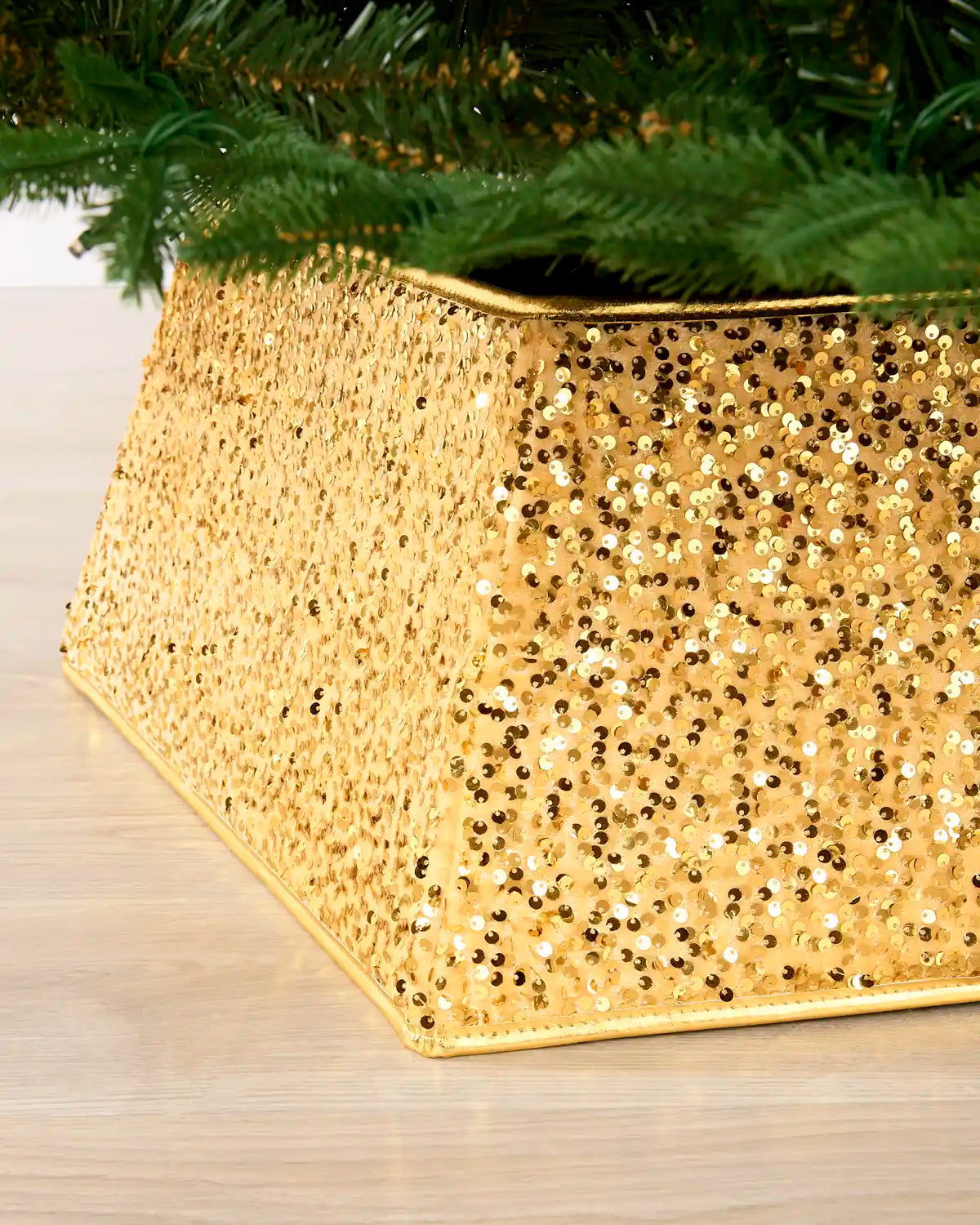 A square gold sequined tree collar with a gold trim, shown holding the green branches of a Christmas tree on a light wood floor.