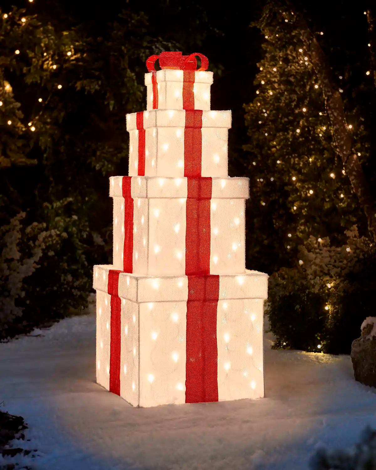 A stacked set of four white gift boxes with red ribbons and glowing warm white string lights, displayed outdoors in the snow against a dark, festive background with Christmas trees.