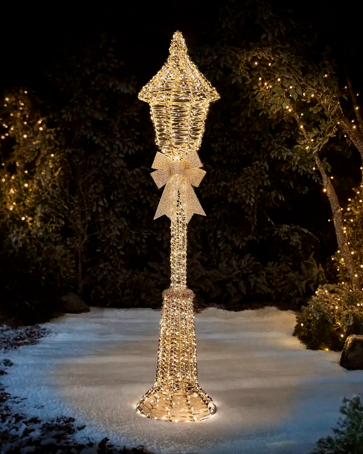 A tall, decorative outdoor light sculpture shaped like a lamp post, covered in warm white lights and accented with a large bow near the top, standing on a snowy path at night with trees in the background also lit with string lights.