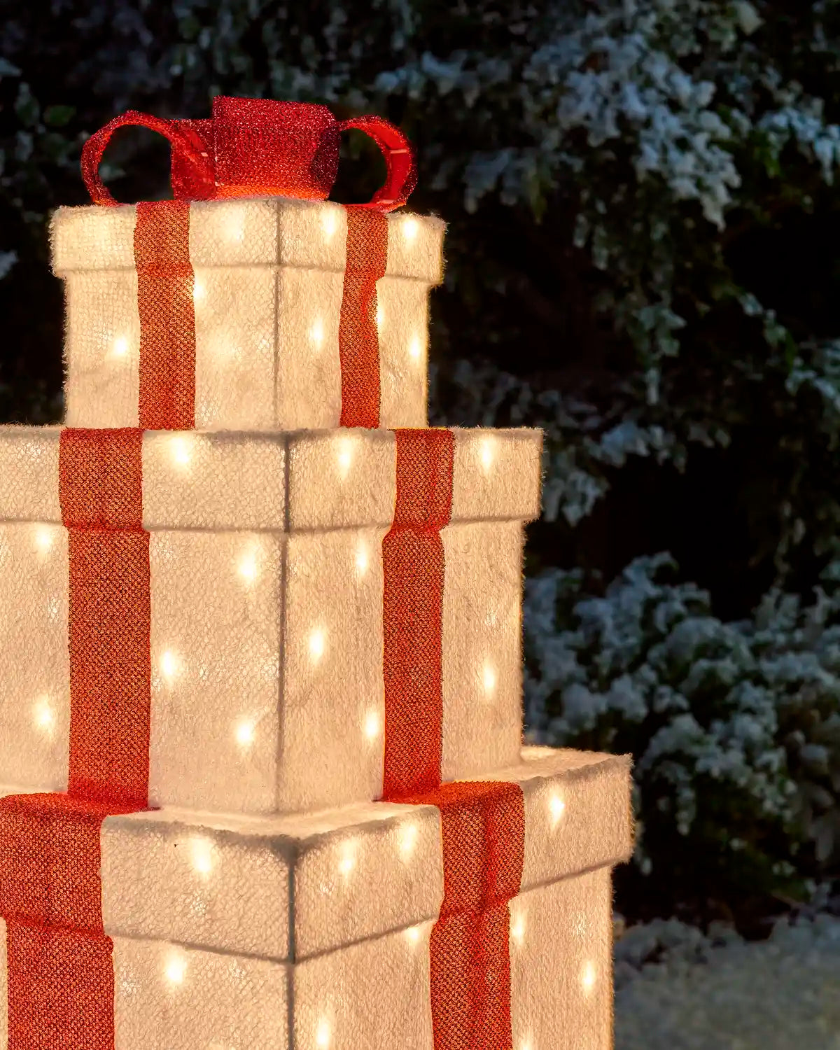 A three-tiered Christmas gift box light decoration with warm white LED lights and red burlap-style ribbons, shown outdoors against a dark, snowy background.