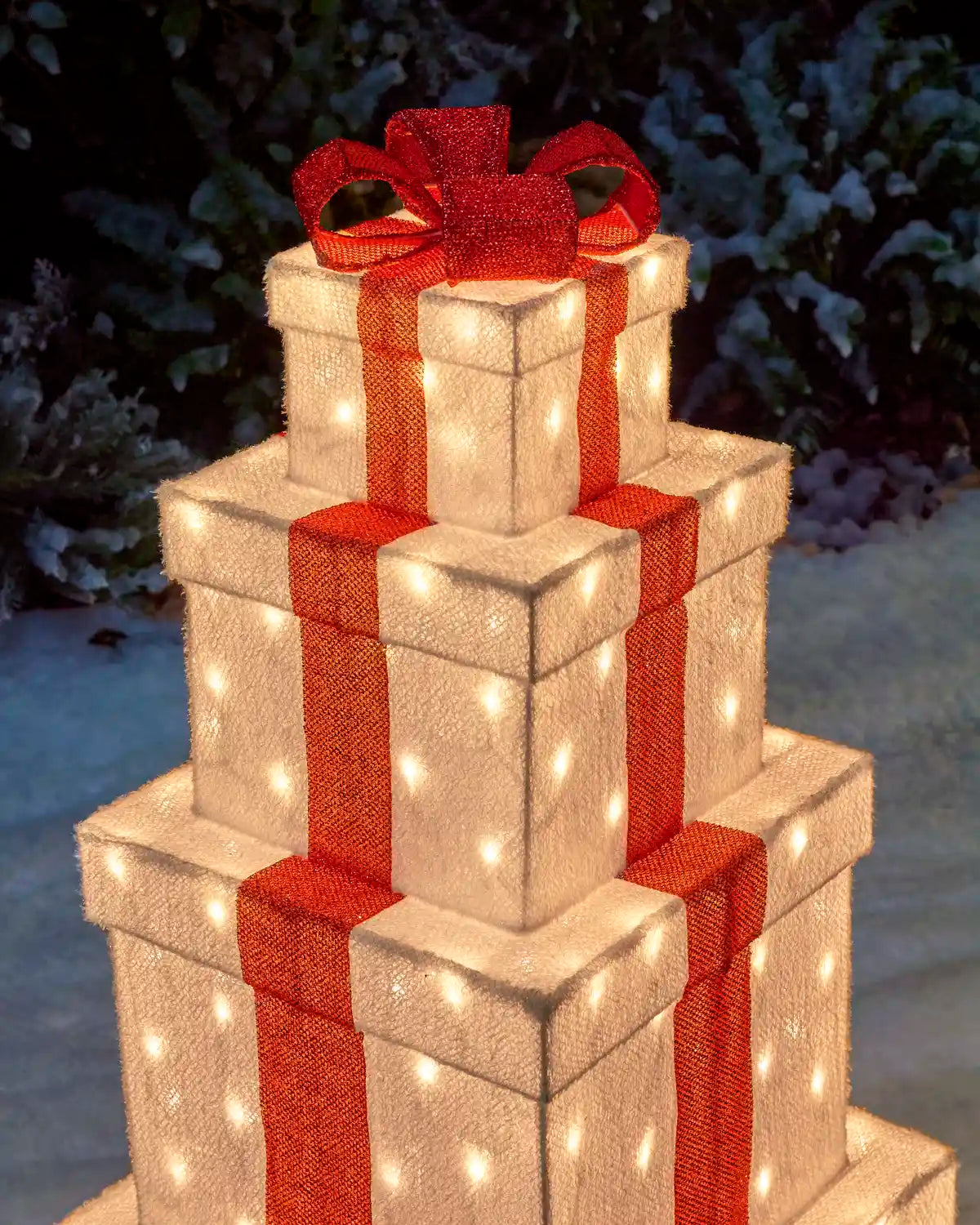 A tiered stack of gift boxes covered in warm white lights, tied with red ribbons and topped with a red bow, set outdoors against a dark snowy background.