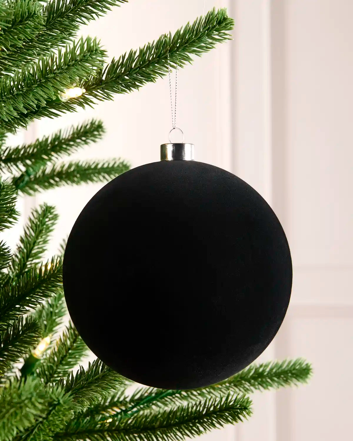 A velvet black Christmas bauble hanging from a green Christmas tree branch with warm string lights, with a light background.