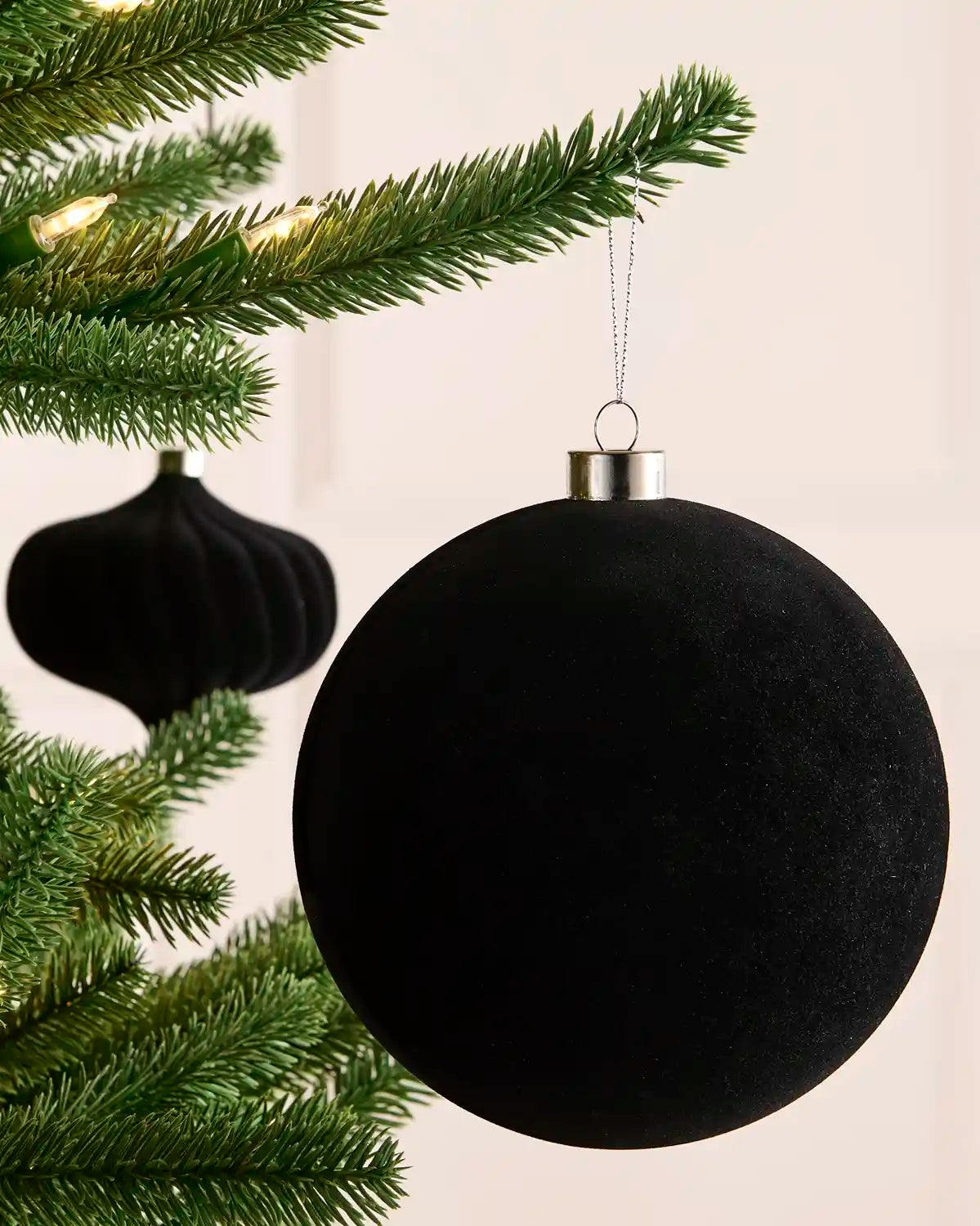A velvet black spherical Christmas ornament hanging from a green Christmas tree branch with warm string lights, with a second black ornament blurred in the background.