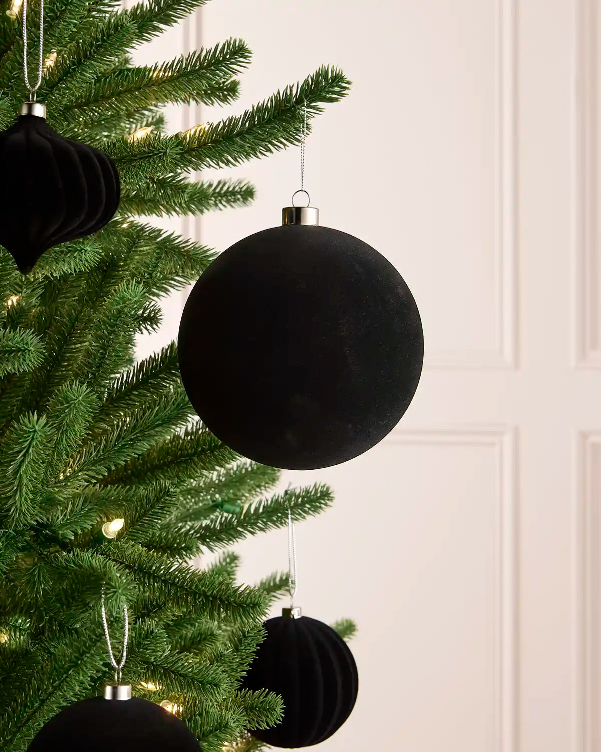 A velvet black spherical Christmas ornament hanging from a green Christmas tree branch with warm string lights, with smaller black ornaments blurred in the background.