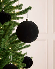 A velvet black spherical Christmas ornament hanging from a green Christmas tree branch with warm string lights, with smaller black ornaments blurred in the background.