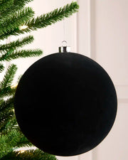 A velvet black spherical Christmas ornament hanging from a silver cap and wire, positioned next to green pine tree branches with warm white string lights against a light cream wall.