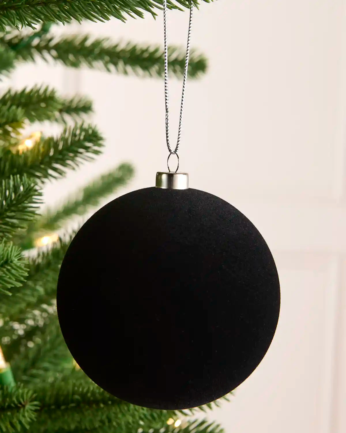 A velvet black spherical Christmas ornament hanging from a silver metal cap on a thin silver string, positioned in front of green pine tree branches with soft warm lights.