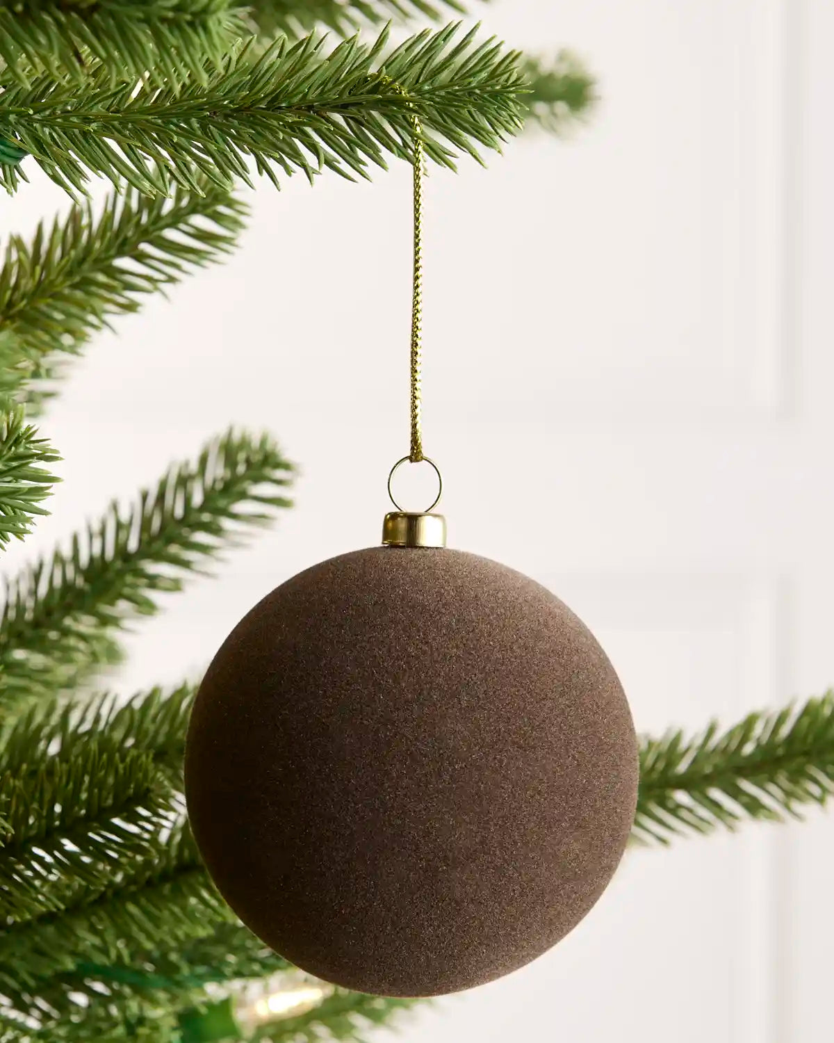 A velvet brown spherical Christmas ornament hanging from a gold-colored wire hanger on a green pine tree branch against a white paneled background.