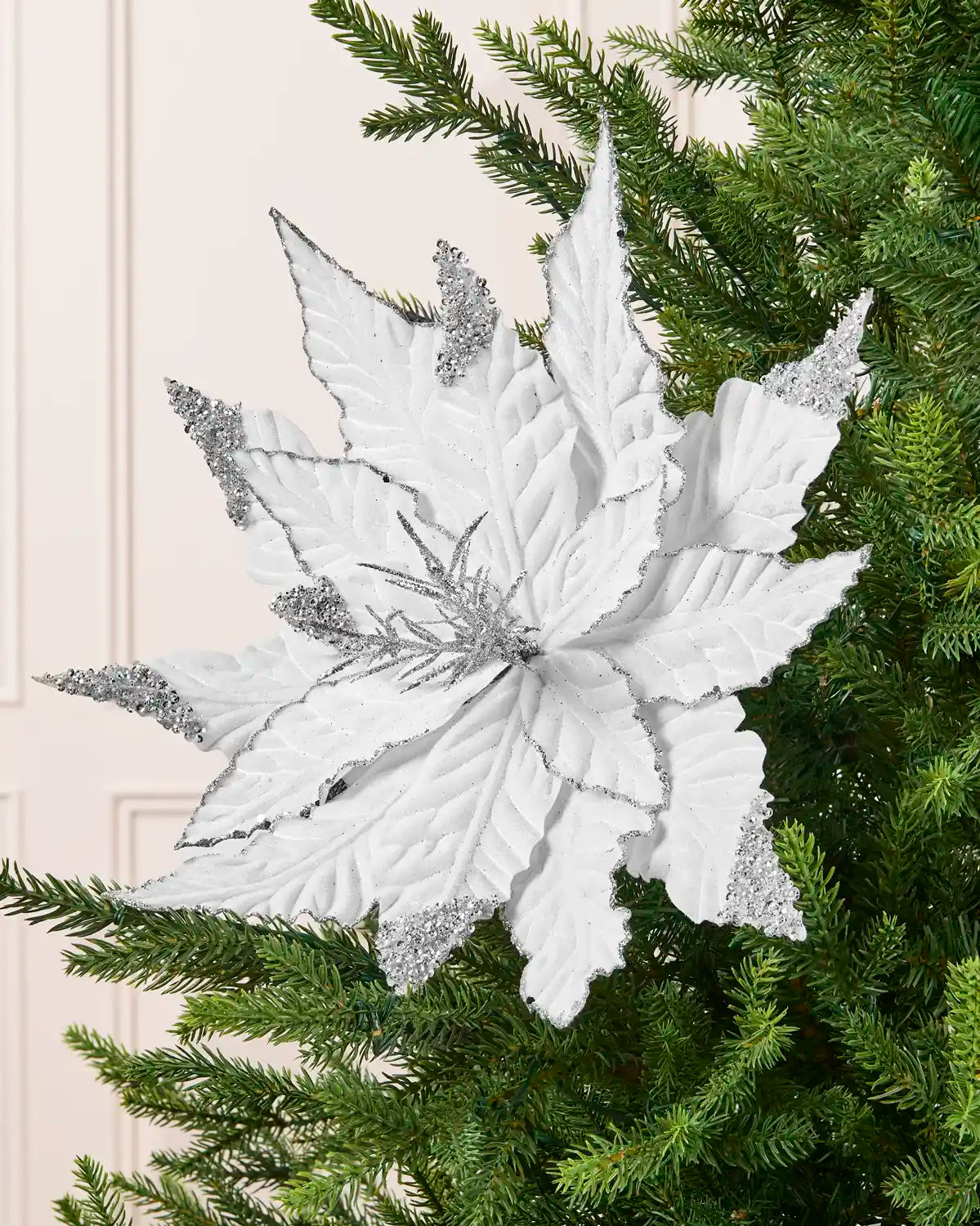 A white poinsettia tree ornament with silver glitter accents, displayed on a green Christmas tree.
