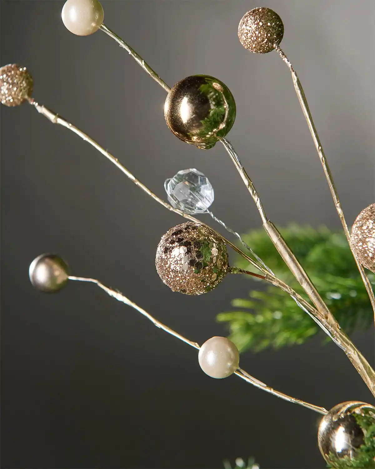 Additional close-up of gold bauble and jewelled spray on tree in front of grey wall background highlighting shine of product