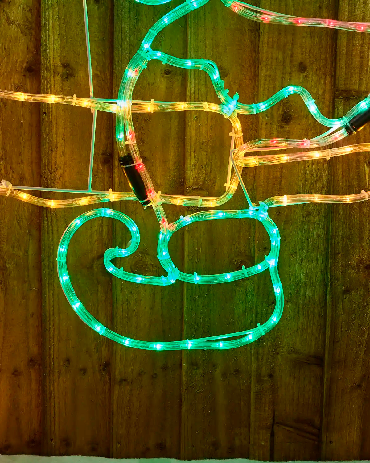 An elves boot LED light Christmas decoration shaped like an elf figure, mounted against a dark wooden fence with a snowy green tree on the right side.
