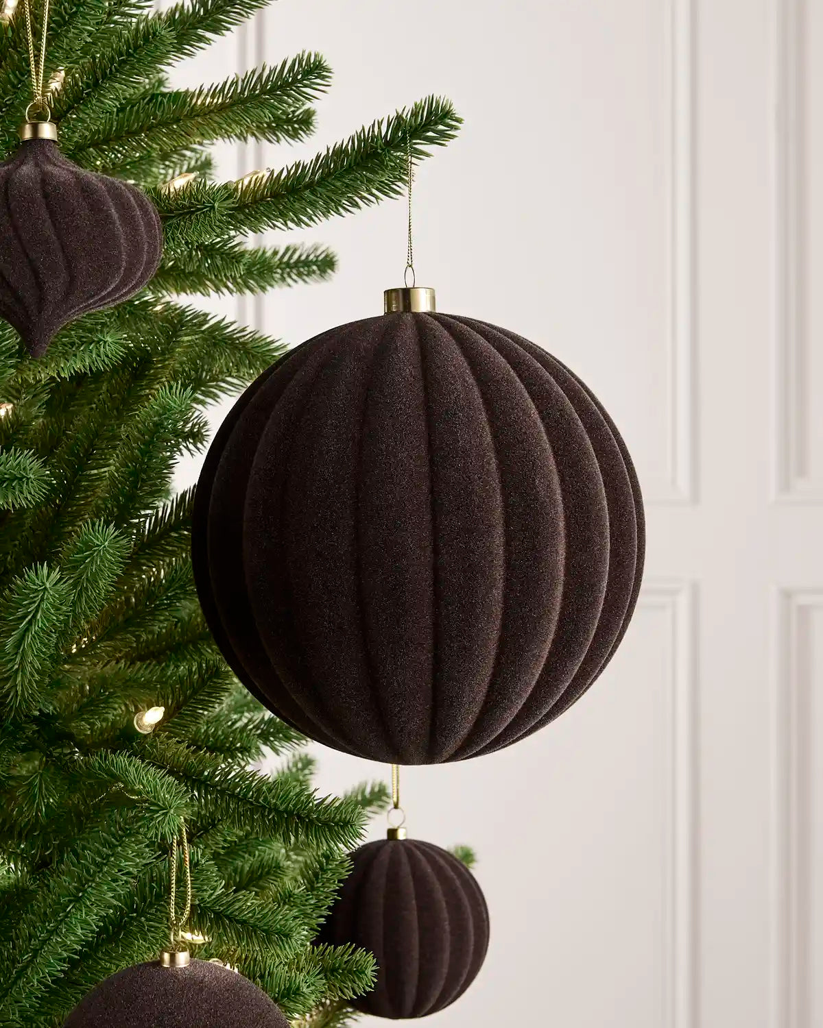 An espresso coloured ribbed fabric Christmas ornament hanging from a green Christmas tree, with other matching ornaments and soft warm lights in the background.