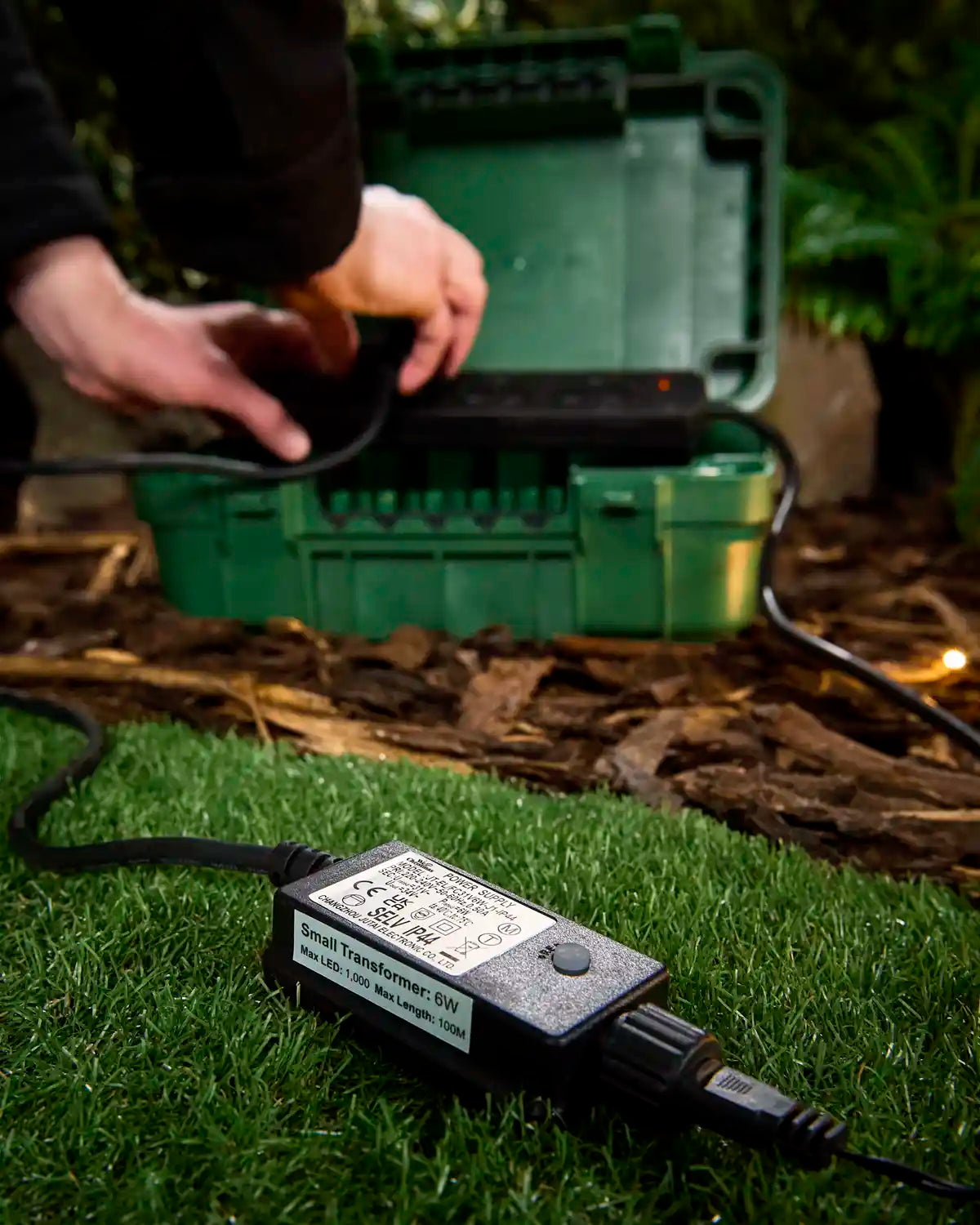 Black 6W transformer for outdoor LED string lights on grass while being connected to a green waterproof storage box.