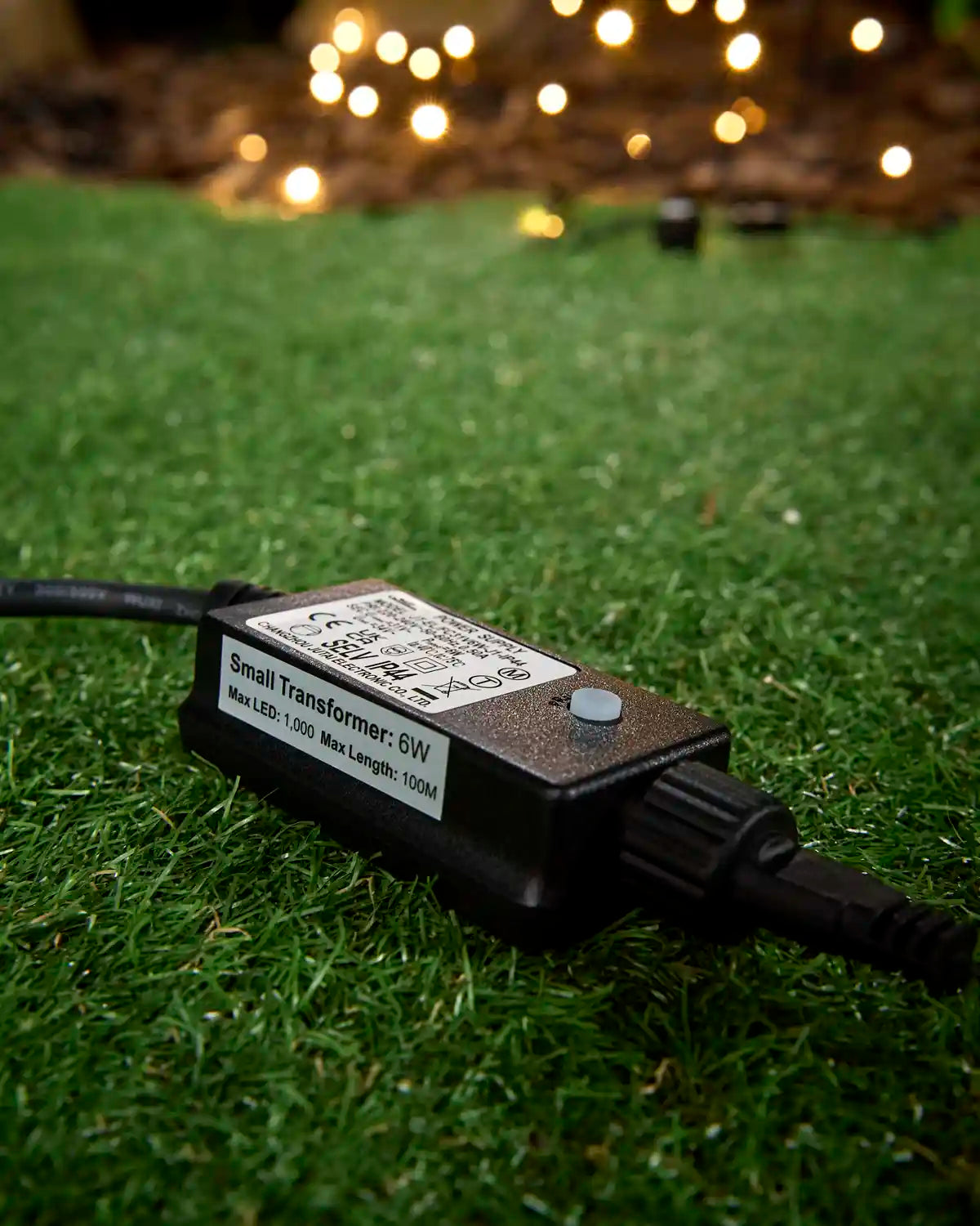 Black 6W transformer for outdoor LED string lights on grass with warm white lights glowing in the background.