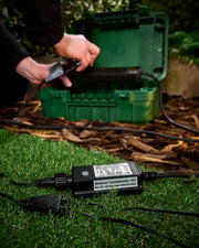 Black 9W transformer for outdoor LED string lights on grass while being connected to a green storage box.