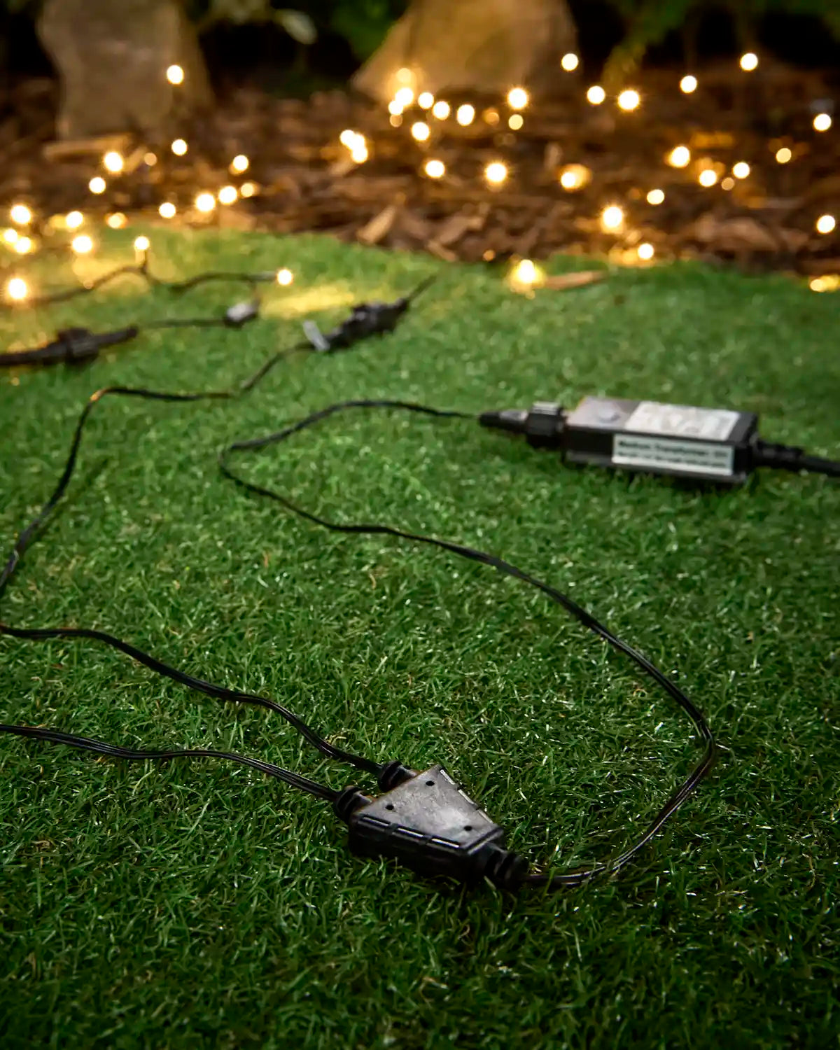 Black 9W transformer with cable splitter for outdoor LED string lights on grass, with warm white lights glowing in the background.