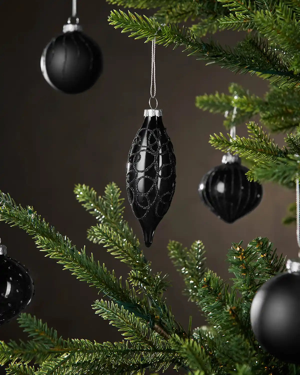 Black Christmas baubles in various shapes hanging on a green tree against a dark background