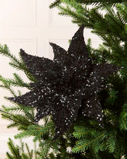 Black Sequin Artificial Poinsettia positioned in Christmas tree branches with a blurry cream background 
