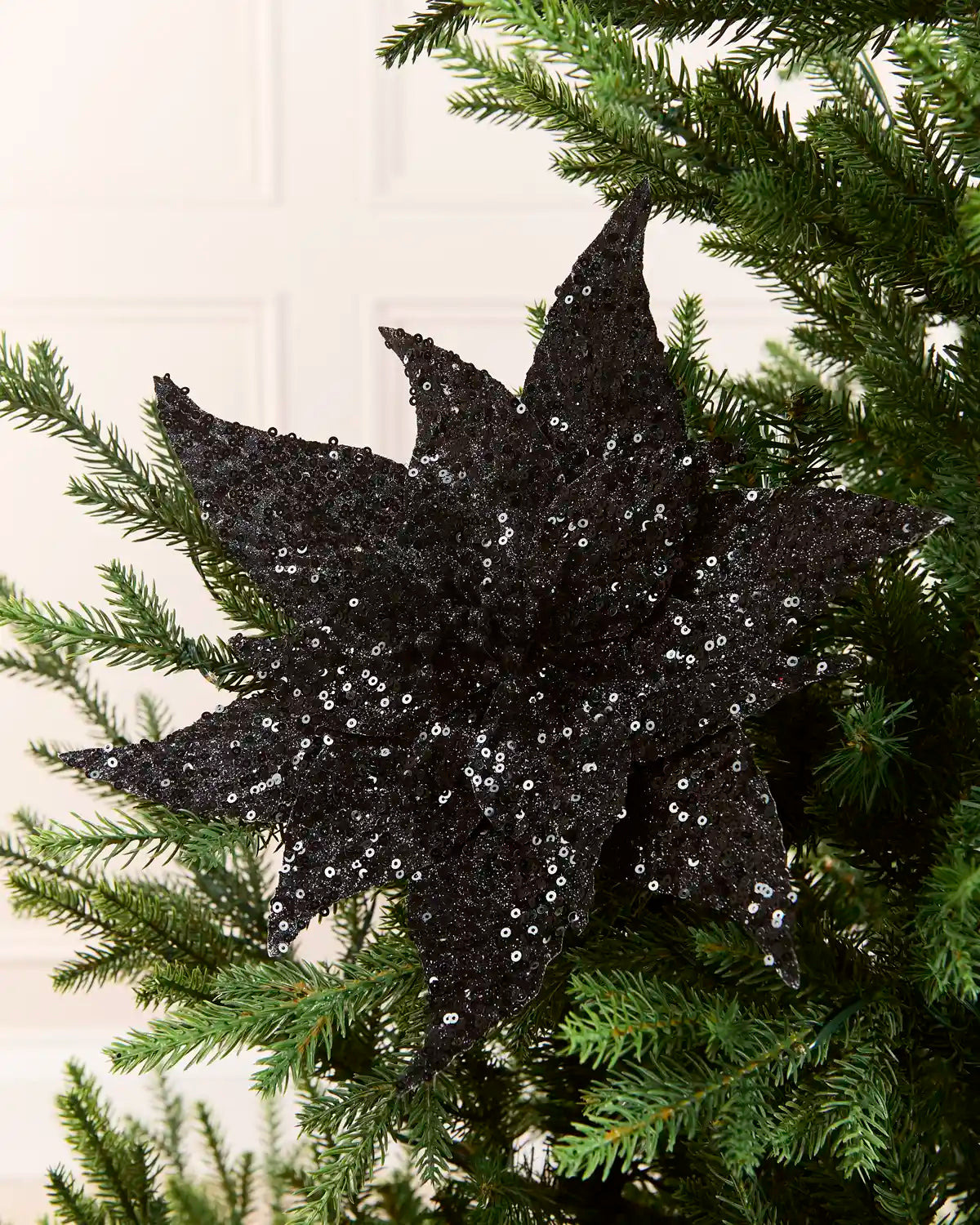 Black Sequin Artificial Poinsettia positioned in Christmas tree branches with a blurry cream background 
