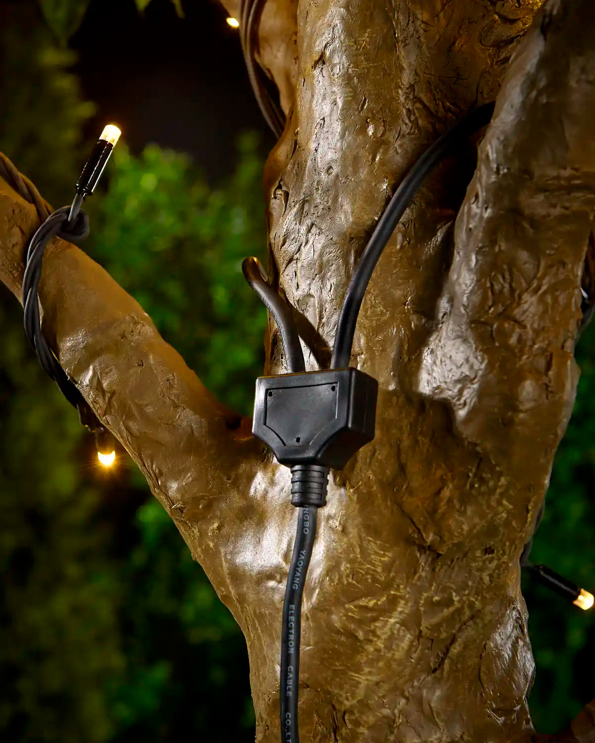 Black Y shaped connector cable attached to a tree with warm white LED string lights and a blurry garden background