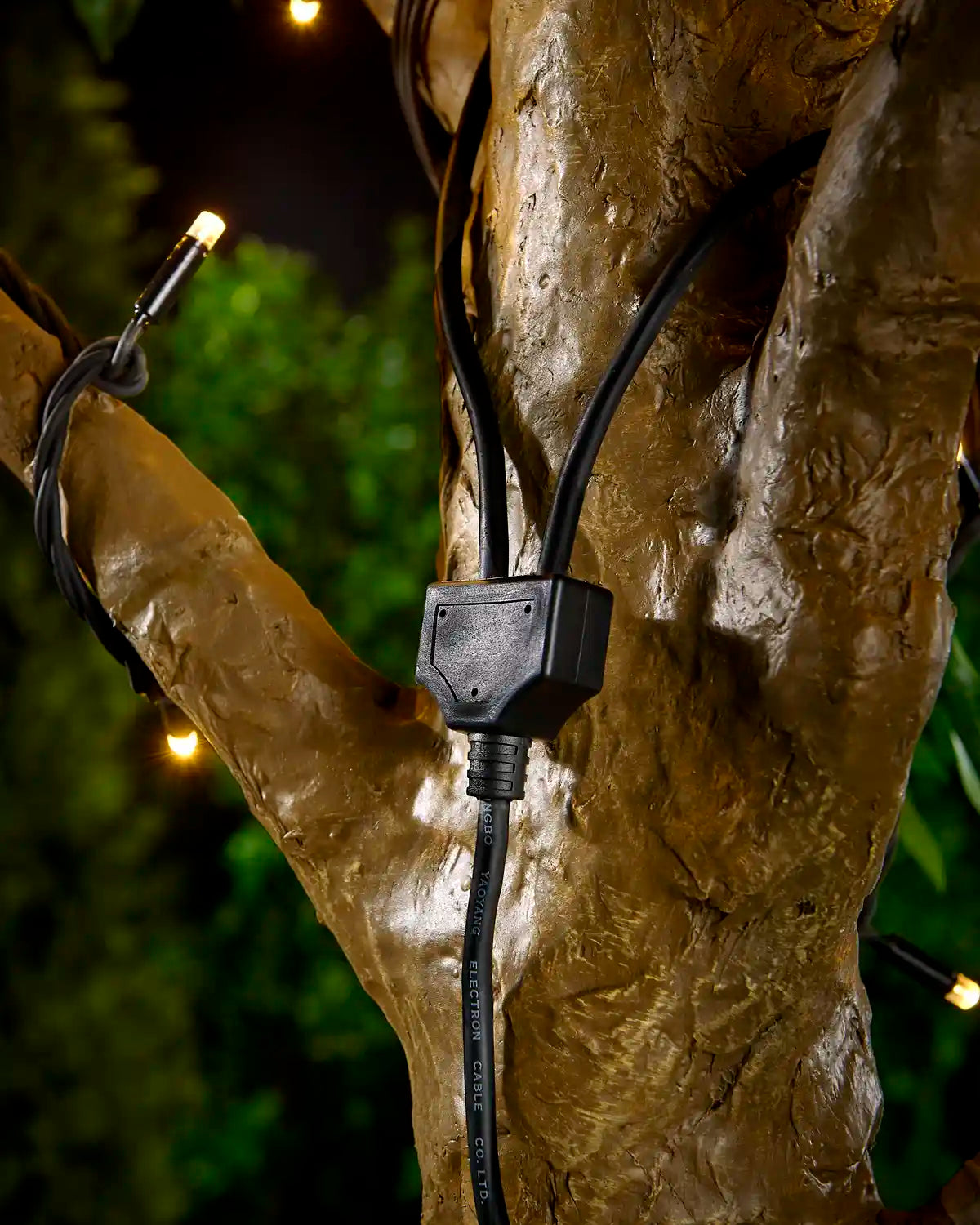 Black Y shaped connector cable attached to tree with blurry garden background and connected to warm white LED string lights