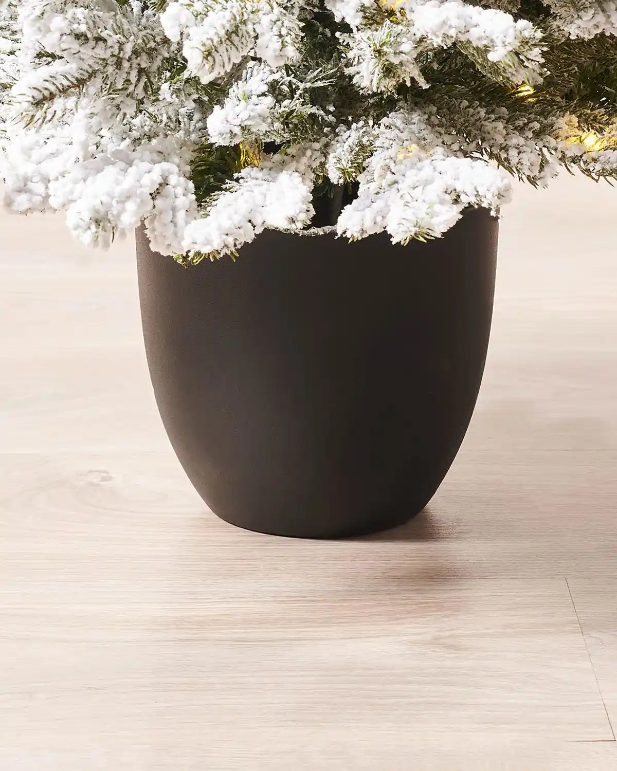 Black ceramic pot with a snow flocked Christmas tree on a light wooden floor