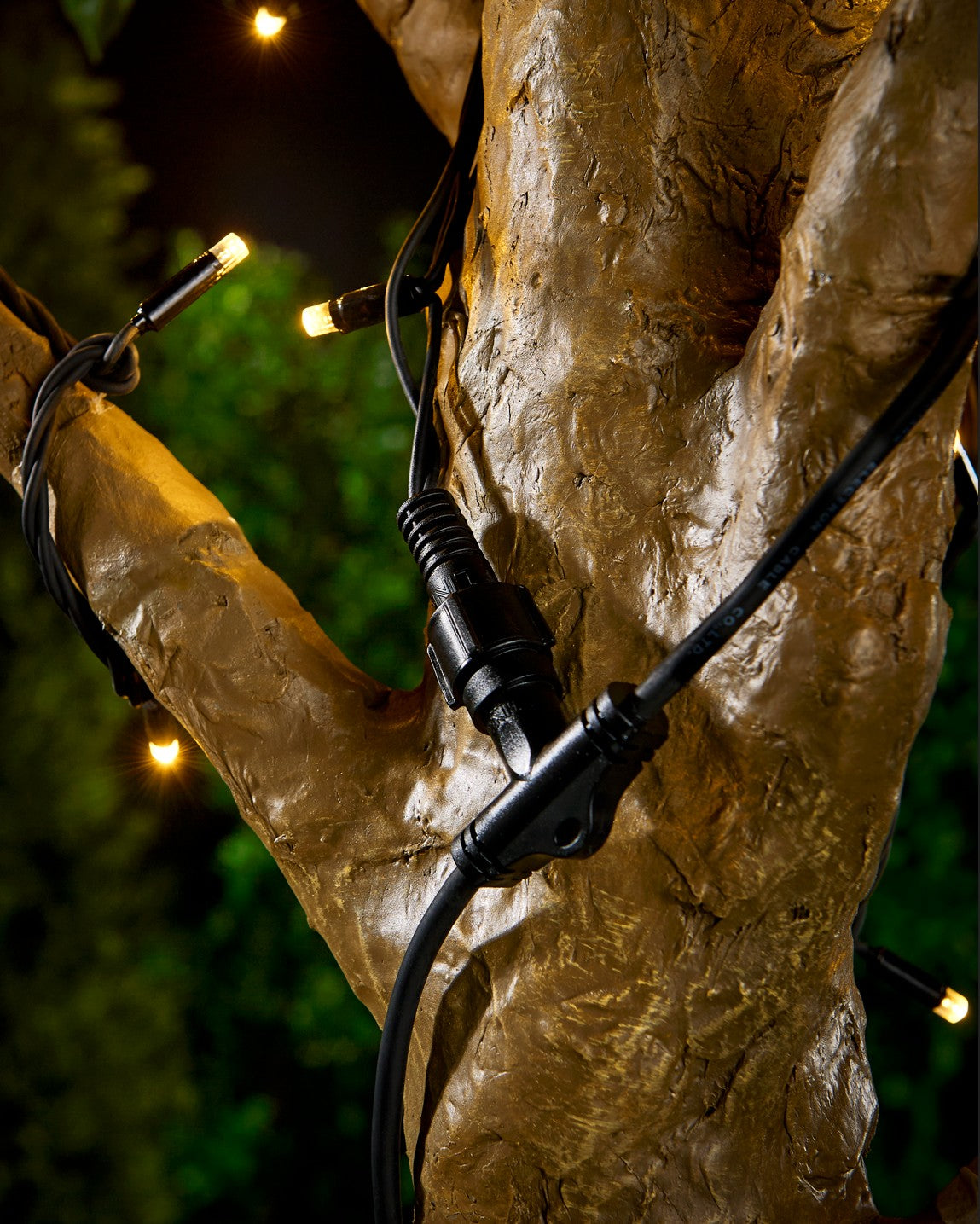 Black connector cable in the shape of a T wrapped around a tree with warm white LED lights and a blurry green background