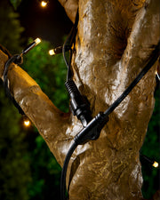 Black connector cable in the shape of a T wrapped around a tree with warm white LED lights and a blurry green background