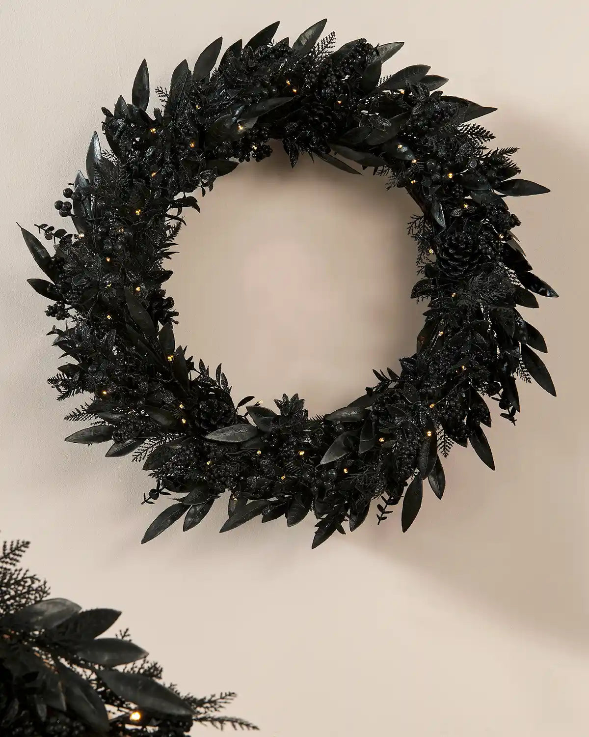 Black decorated wreath with black pine cones, berries and warm white lights against a cream wall