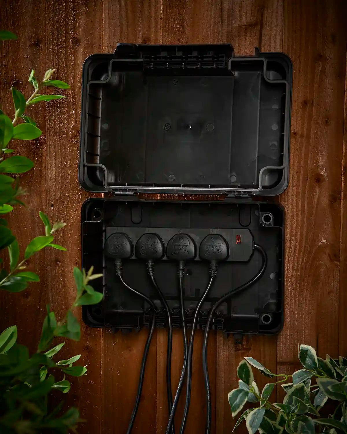 Black electrical junction box with wires on a wooden fence, surrounded by green plants.