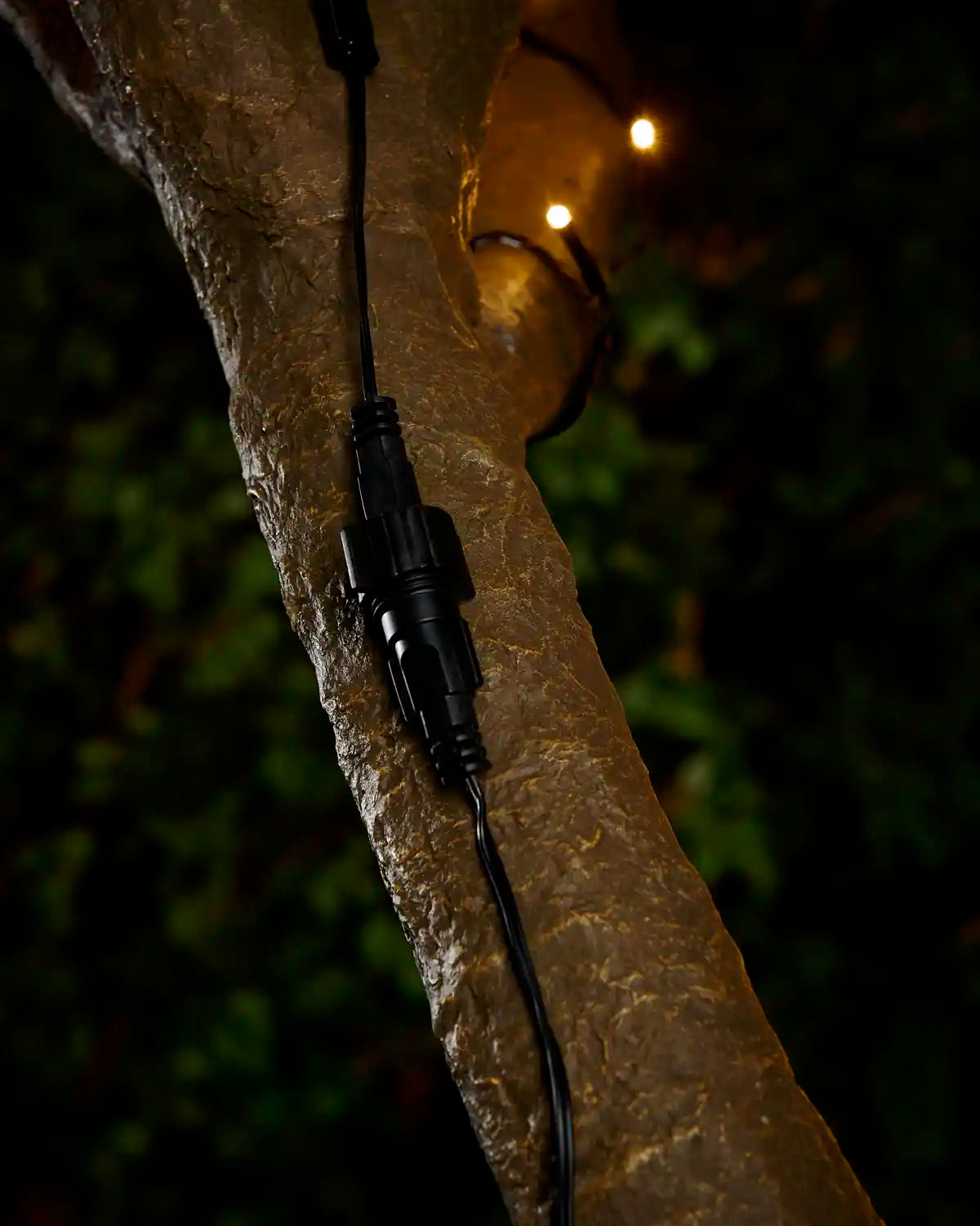 Black extension cable connection point with screw cap attached to tree with blurry LEDs in the background. 