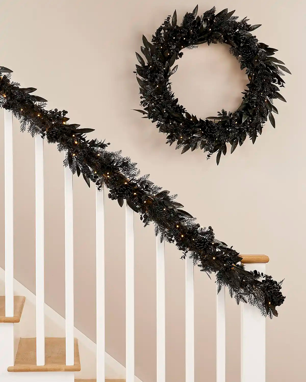 Black garland and matching wreath with warm white lights on a stair case banister against a cream wall